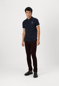 Mornarska polo majica s kratkim tamnosivim akcentima, s izvezenim logom na prsima. Uparena s tamnocrvenim kordura hlačama i crnim cipelama.