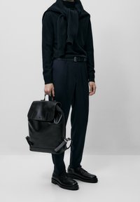 Zwarte leren rugzak met een klep en verstelbare bandjes, gedragen door een persoon in donkere kleding, staand tegen een neutrale achtergrond.