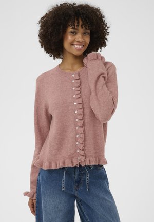 Ung kvinde med krøllet hår iført lyserød, flæset knapcardigan og blå jeans, smilende med hånden tæt på ansigtet.