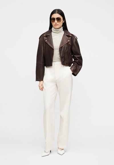 Γυναίκα που φοράει σκούρο καφέ κοντό δερμάτινο μπουφάν, ριγέ ζιβάγκο, λευκό παντελόνι με ψηλή μέση, άσπρες γόβες και γυαλιά ηλίου, στέκεται με το ένα χέρι στην τσέπη.