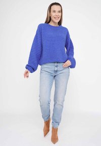 Blauer, gerippter Strickpullover mit tiefen Schultern, langen Ärmeln und cropped Passform. Getragen mit hellblauen Jeans und beigen spitzen Ankle Boots.