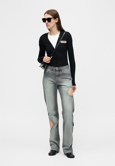 Femme portant un haut à manches longues noir avec un sous-shirt blanc, un jean gris avec des trous aux jambes, des bottes noires, des lunettes de soleil noires et un petit sac bandoulière noir.