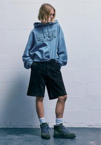 Világoskék kapucnis pulcsi "STRANGER THINGS" grafikával, fekete velúr rövidnadrág, zöld túracipők kék fűzős, és fehér csíkos zoknik.