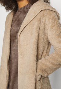 Person iført en beige fuzzy frakke over en brun ternet top med høj krave, med den ene hånd i frakkens lommer.