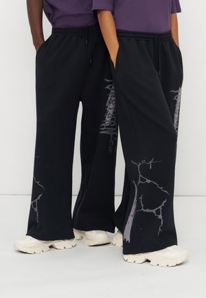 Deux personnes portant un pantalon noir à jambes larges avec des motifs gris ressemblant à des fissures et des baskets blanches épaisses, debout côte à côte.