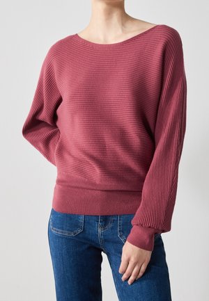 Persona con un suéter malva de manga larga con textura acanalada y un cuello ancho, y vaqueros azules, con la mano apoyada cerca de la cadera.