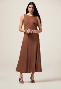 Robe maxi sans manches marron en tissu texturé avec un corsage ajusté. Des accessoires en métal doré complètent le look, associés à des chaussures ouvertes.
