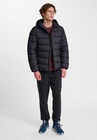 Zwarte donsjas met capuchon, doorgestikt ontwerp en ritssluiting aan de voorkant, gecombineerd met een donkere broek en blauwe sportschoenen.