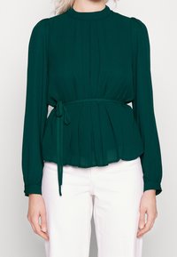Blusa verde plissettata con collo alto, maniche lunghe e cintura annodata in vita, realizzata in tessuto leggero; abbinata a pantaloni bianchi.