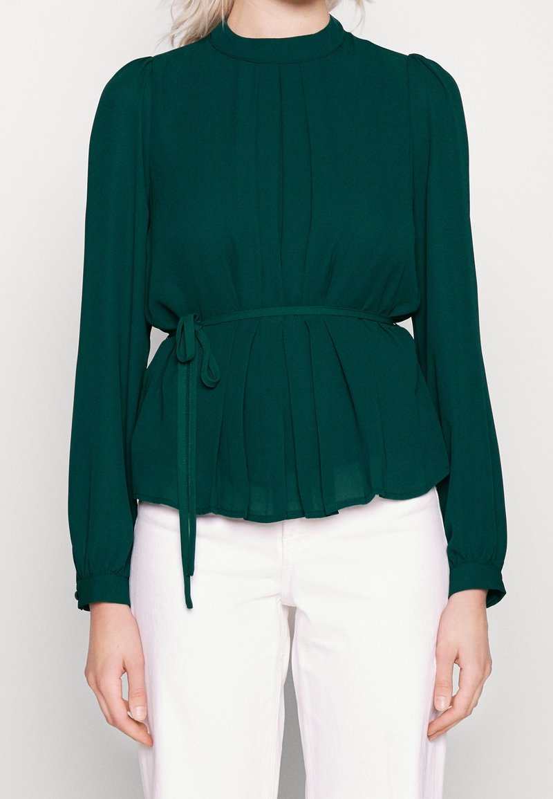 Blusa verde plissettata con collo alto, maniche lunghe e cintura annodata in vita, realizzata in tessuto leggero; abbinata a pantaloni bianchi.