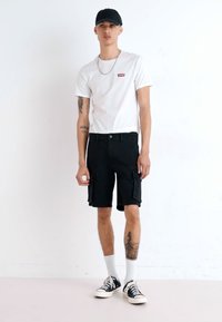 Fehér rövid ujjú póló piros logóval, fekete cargo rövidnadrág oldalzsebekkel, fekete sapka, fehér crew zokni és fekete vászon sportcipő.