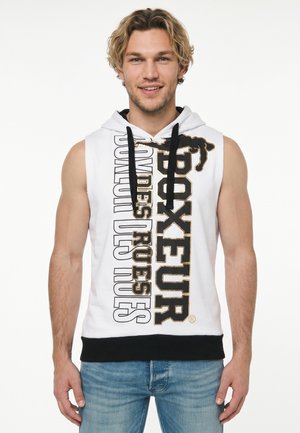 Mann mit blondem Haar, der einen weißen ärmellosen Hoodie mit schwarzen Kordeln und auffälligem schwarz-goldenem Schriftzug trägt, steht vor weißem Hintergrund.