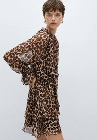 Haljina od prozirnog materijala s uzorkom leopard, s volanima na rukavima i slojevitim rubom. Zemljane tonove smeđe i bež boje čine lagani, lepršavi dizajn.