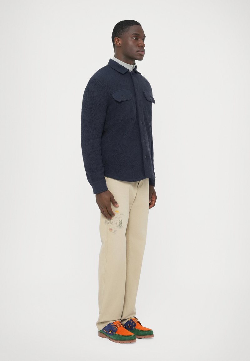 Navy fleece shirt jacket with two front pockets, beige trousers featuring embroidery, and multicolored sneakers with layered textures and colors.