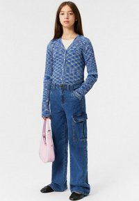 Blå texturerad cardigan med V-ringning, tillsammans med högmidjade blå cargobyxor med sidofickor. Håller en ljusrosa handväska.