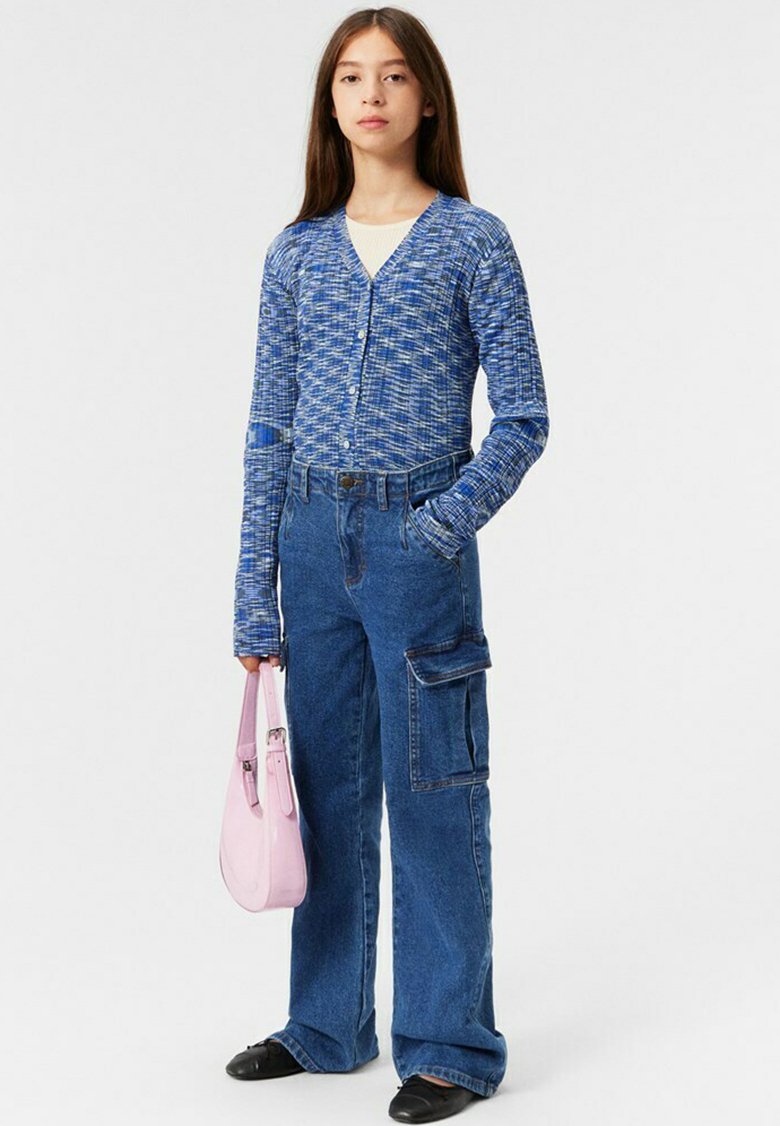 Blå texturerad cardigan med V-ringning, tillsammans med högmidjade blå cargobyxor med sidofickor. Håller en ljusrosa handväska.