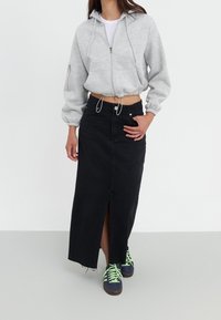 Kobieta w jasnoszarej, krótkiej, zapinanej na zamek bluzie z kapturem, czarnej, wysokiej spódnicy maxi z rozcięciem z przodu oraz granatowych trampkach z neonowymi zielonymi sznurowadłami.