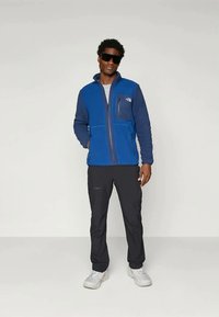 Kék polárdzseki, elöl cipzárral, bal mellkasi zsebbel, fekete nadrággal és fehér sportcipővel viselve. A modell semleges háttér előtt áll.