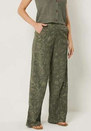 Kvinde iført olivengrønne bukser med vide ben og beige blomstret broderi samt en ærmeløs, strikket vest med knapper, hånd i lommen.