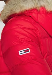 Rød pufferjakke med hætte kantet med pels. Har et quiltet design og et lille broderet logo på ærmet. Blød tekstur.