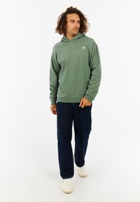 Groene hoodie met een capuchon en een klein embleem op de borst. Gecombineerd met donkerblauwe jeans en witte sneakers. Glad stoftextuur.