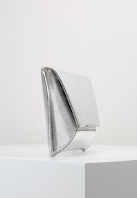 Silverfärgad metalliskt clutch-väska med en slät yta, triangulär form och flikstängning. Har en glansig finish och minimalistisk design.