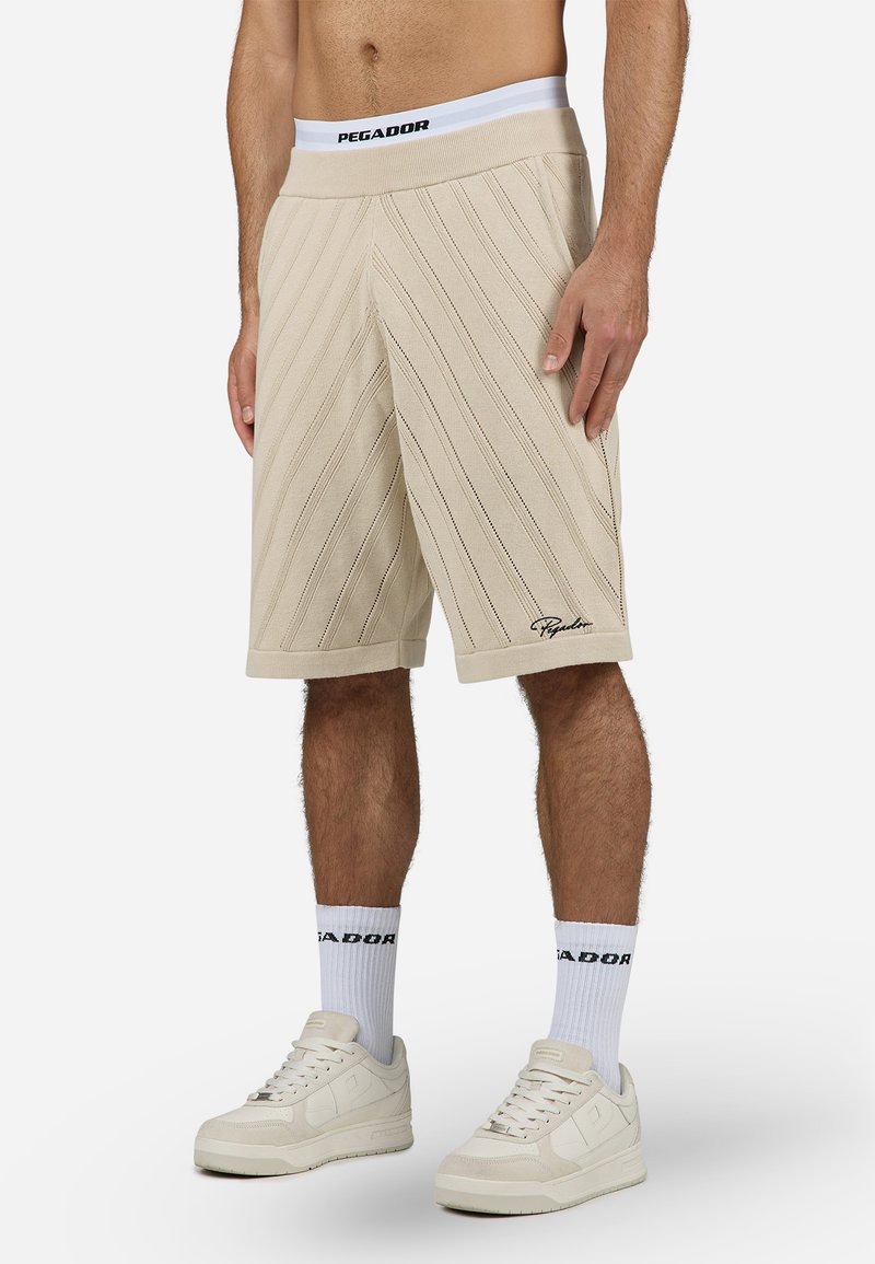 Shorts beiges avec des rayures texturées diagonales, taille élastique avec la marque "PEGADOR", longueur genoux, associés à des chaussettes blanches et des baskets.