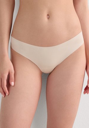 Kvinde iført sømløst beige bikini-inspireret undertøj, der viser mave og øvre lår mod en ensartet lys baggrund.
