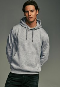 Gray hoodie made of soft fabric with a kangaroo pocket, drawstrings, and ribbed cuffs. Worn over dark jeans against a neutral backdrop.