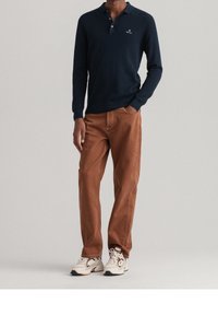 Marineblaues Langarm-Polo-Shirt mit Kragen und Logo, kombiniert mit braunen Hosen und weißen Sneakern mit schwarzen Akzenten. Flache Textur, lässige Passform.