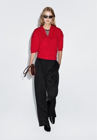 Rode cropped trui met een V-hals, gecombineerd met zwarte wijde broek. Het model houdt een bruine handtas vast en draagt zwarte zonnebril.