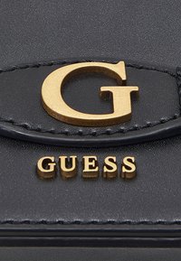 Černá peněženka z polyesterového materiálu s výrazným zlatým logem "G" a raženým textem "GUESS". Hladký povrch s viditelnými prošívacími detaily.