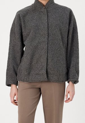 Personne portant une veste à fermeture éclair ample et texturée de couleur gris foncé avec un col montant, ainsi qu'un pantalon beige, sur un fond blanc uni.