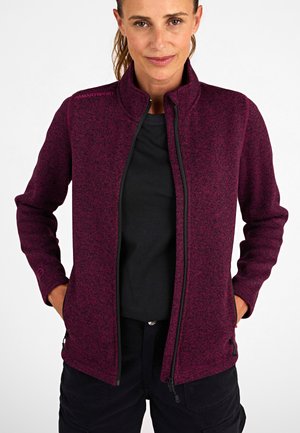 Vrouw in een donkerpaarse fleece met ritssluiting over een zwart shirt, met de handen in de zakken, staand tegen een plain witte achtergrond.
