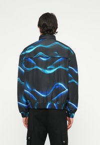 Čierna vetrovka so vzormi modrých vĺn, elastický pás a manžety. Obsahuje ventilačné otvory vzadu a je vyrobená z ľahkého materiálu.
