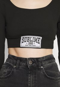Black long-sleeve cropped top with a white "SUBLIME 1995 NEW YORK STATE" patch; paired with high-waisted dark denim jeans.