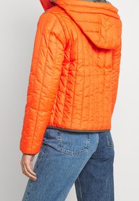 Osoba v jasně oranžové prošívané bundě s kapucí a modrých džínách, zobrazená zezadu na jednoduchém pozadí.