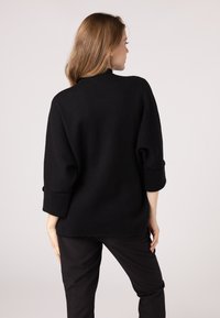 Maglione nero oversize con maniche arrotolate a 3/4, colletto alto e una texture morbida, abbinato a pantaloni neri.