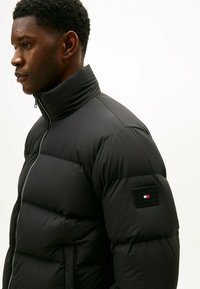 Giacca a vento nera con colletto alto e design trapuntato. Presenta una patch con logo sul braccio e una zip frontale. Texture morbida e isolata.