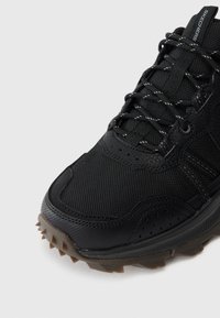 Bottine de sport noire avec tige en mesh texturé, bout renforcé et semelle en caoutchouc avec des pointes pour l'adhérence. Les lacets sont réfléchissants.