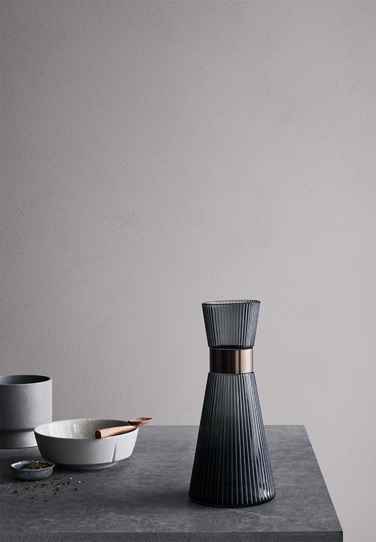 Carafas de vidrio con textura acanalada vertical, color gris oscuro, banda de metal en el centro, colocada sobre una superficie de piedra oscura junto a cuencos.