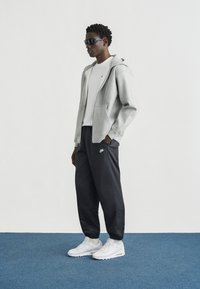 Γκρι φούτερ με κουκούλα και μπροστινές τσέπες, λευκό μπλουζάκι, μαύροι παντελόνες joggers με λογότυπο Nike και λευκά αθλητικά παπούτσια. Το μοντέλο στέκεται σε μπλε χαλί μπροστά από άσπρο φόντο.