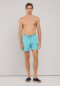 Pantaloni da bagno turchesi con cintura elastica e dettagli con cordino. Il modello è a piedi nudi, mostrando un design minimalista.