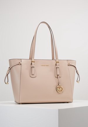 Sac cabas beige Michael Kors avec doubles bretelles, pendentif logo doré et détails de boucle latérale sur une surface blanche avec un fond uni.