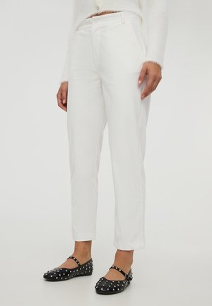 Persona che indossa pantaloni bianchi a coste, un maglione bianco soffice e scarpe Mary Jane nere con borchie, in piedi su uno sfondo neutro.