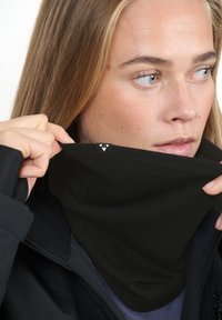 Svart halsvärmare i mjukt tyg, med sydd kant och en liten logotypetikett. Modellens hand justerar halsvärmaren.