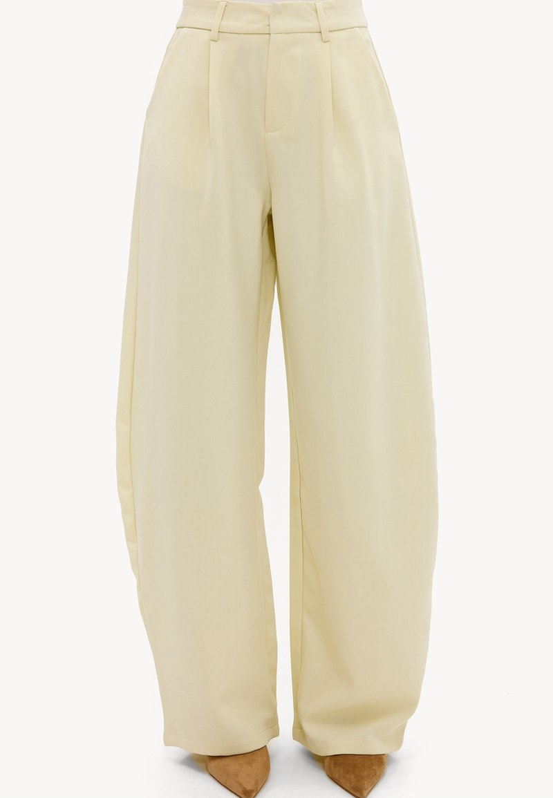 Pantalon large beige taille haute avec plis et passants de ceinture, porté avec des chaussures pointues en daim marron.