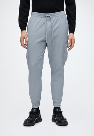 Pantalon de jogging gris clair avec taille à cordon et poches latérales, porté avec des baskets noires et un haut noir à manches longues.