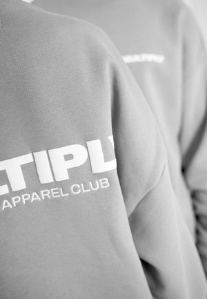 Gray sweatshirt featuring bold white embroidered text that reads "MULTIPLY APPAREL CLUB." The fabric appears soft with a smooth texture.