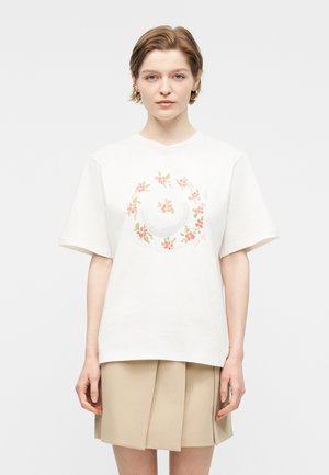 Γυναίκα με κοντά μαλλιά φοράει λευκό υπερμεγέθες μπλουζάκι με σχέδιο στεφανιού με λουλούδια και μπεζ πλισέ φούστα, στέκεται μπροστά σε απλό φόντο.